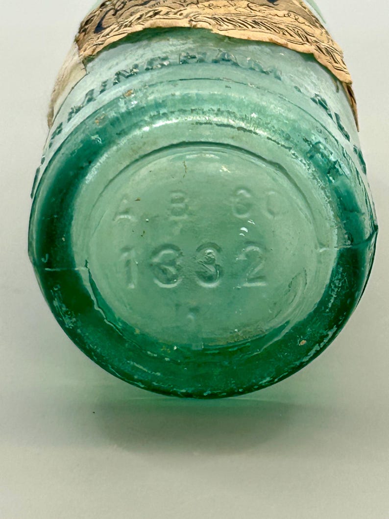 Pode incluir: Uma garrafa de vidro verde-claro vintage com um r&oacute;tulo de papel bege parcial enrolado no gargalo. A base circular apresenta um texto em relevo com A B CO e o n&uacute;mero 1332. O vidro &eacute; transl&uacute;cido com bolhas de ar vis&iacute;veis no material.