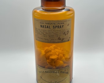 Pode incluir: Uma garrafa de vidro âmbar rotulada como "Nasal Spray" com uma rolha de cortiça. O rótulo indica o conteúdo de 500 comprimidos e lista os ingredientes. A garrafa está cheia de comprimidos laranja e tem um rótulo da The S.E. Massengill Company.
