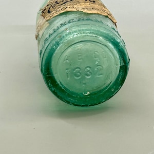 Pode incluir: Uma garrafa de vidro verde-claro vintage da Coca-Cola com um r&oacute;tulo de papel desgastado apresentando o logotipo da Coca-Cola. A base circular da garrafa est&aacute; gravada com as letras AB CO e o n&uacute;mero 1332, mostrando claramente a parte inferior.