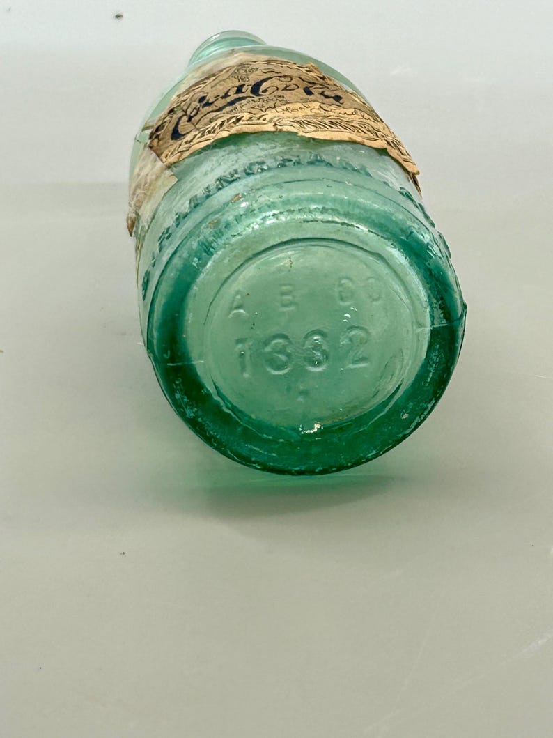 Pode incluir: Uma garrafa de vidro verde-claro vintage da Coca-Cola com um r&oacute;tulo de papel desgastado apresentando o logotipo da Coca-Cola. A base circular da garrafa est&aacute; gravada com as letras AB CO e o n&uacute;mero 1332, mostrando claramente a parte inferior.