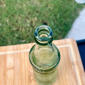May include: A small, empty, clear green glass bottle with a narrow neck and a rounded body. The bottle features a thick, circular rim at the top opening and a smooth, transparent surface that allows light to pass through the glass structure.
