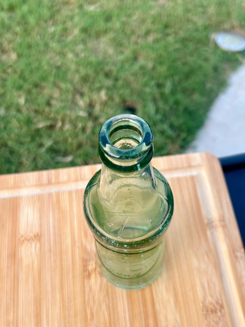 May include: A small, empty, clear green glass bottle with a narrow neck and a rounded body. The bottle features a thick, circular rim at the top opening and a smooth, transparent surface that allows light to pass through the glass structure.