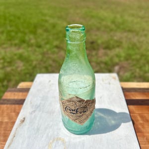 Pode incluir: Uma garrafa de Coca-Cola vintage de vidro verde claro com gargalo estreito e um r&oacute;tulo de papel desgastado em forma de diamante com o log&oacute;tipo da Coca-Cola em escrita preta. A garrafa tem uma base arredondada e uma superf&iacute;cie lisa e transl&uacute;cida.