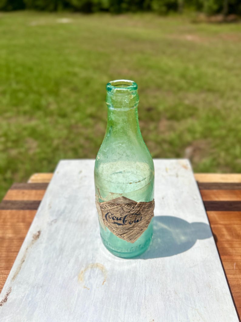Pode incluir: Uma garrafa de Coca-Cola vintage de vidro verde claro com gargalo estreito e um r&oacute;tulo de papel desgastado em forma de diamante com o log&oacute;tipo da Coca-Cola em escrita preta. A garrafa tem uma base arredondada e uma superf&iacute;cie lisa e transl&uacute;cida.