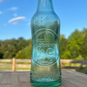 May include: A vintage light green glass soda bottle with an embossed circular logo featuring the text The Original 3 Cent Drink. The base of the bottle includes embossed text reading Trademark Reg U.S. Pat Off and Contents 6 Fl. Oz.