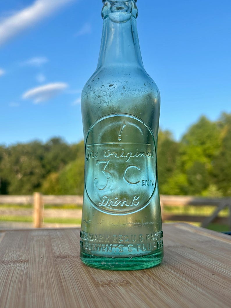 May include: A vintage light green glass soda bottle with an embossed circular logo featuring the text The Original 3 Cent Drink. The base of the bottle includes embossed text reading Trademark Reg U.S. Pat Off and Contents 6 Fl. Oz.