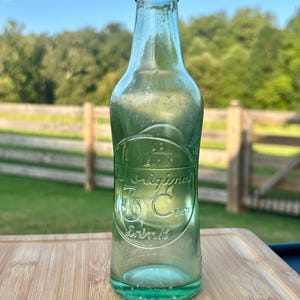 May include: A vintage light green glass soda bottle with a narrow neck and a circular embossed logo on the front. The embossed text reads The Original 5 Cent Drink. The bottle has a smooth, rounded body and a clear, translucent appearance.