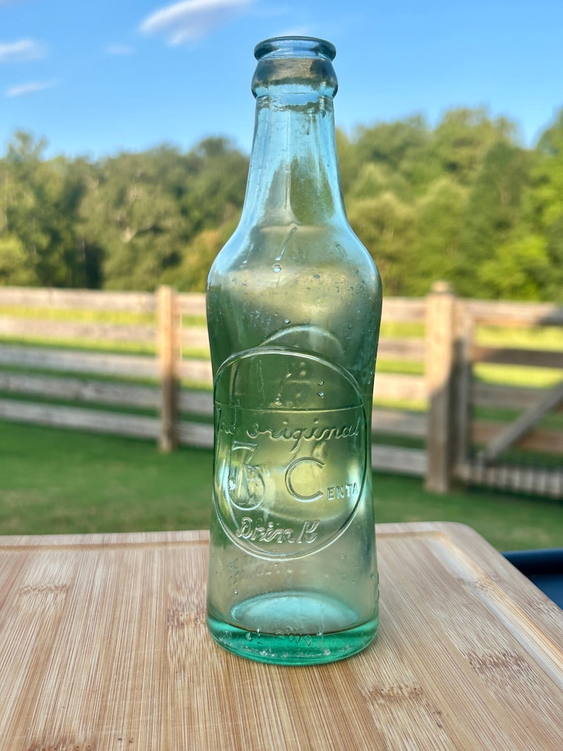May include: A vintage light green glass soda bottle with a narrow neck and a circular embossed logo on the front. The embossed text reads The Original 5 Cent Drink. The bottle has a smooth, rounded body and a clear, translucent appearance.