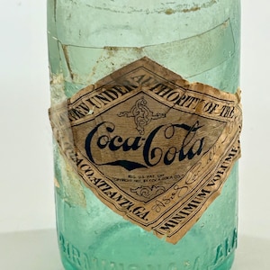 Pode incluir: Uma garrafa de vidro verde claro vintage da Coca-Cola com um r&oacute;tulo de papel em forma de diamante com texto preto que diz Bottled Under Authority of the Coca-Cola Co. Atlanta, GA. A base de vidro tem gravada a palavra Birmingham, ALA.