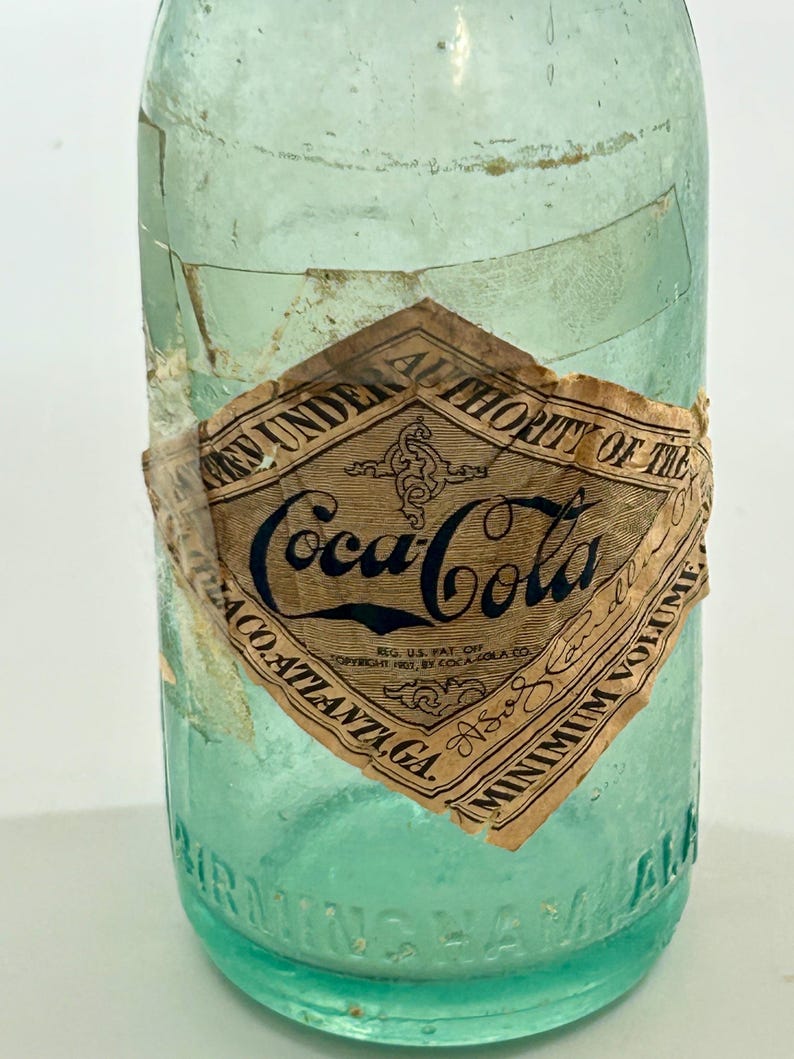 Pode incluir: Uma garrafa de vidro verde claro vintage da Coca-Cola com um r&oacute;tulo de papel em forma de diamante com texto preto que diz Bottled Under Authority of the Coca-Cola Co. Atlanta, GA. A base de vidro tem gravada a palavra Birmingham, ALA.