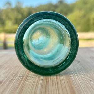 May include: A circular view of the bottom of a dark green glass bottle. The base is thick and translucent, showing a concave center with a smooth, rounded texture. The glass has a deep emerald hue that transitions to a lighter, clearer center.