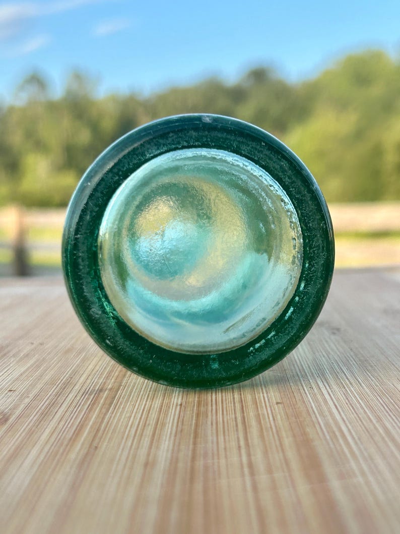 May include: A circular view of the bottom of a dark green glass bottle. The base is thick and translucent, showing a concave center with a smooth, rounded texture. The glass has a deep emerald hue that transitions to a lighter, clearer center.