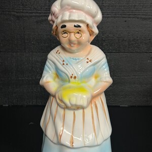 May include: A ceramic figurine of a woman wearing a white chef's hat and apron, holding a yellow loaf of bread. The figurine has gold-rimmed glasses and a blue and white dress. The overall design is vintage and charming.