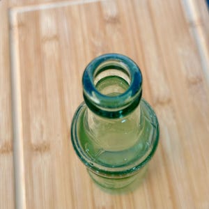 May include: A small, translucent green glass bottle viewed from directly above. The bottle features a rounded neck and a thick, circular rim opening. The glass has a smooth, reflective surface with subtle variations in the green tint throughout the body.