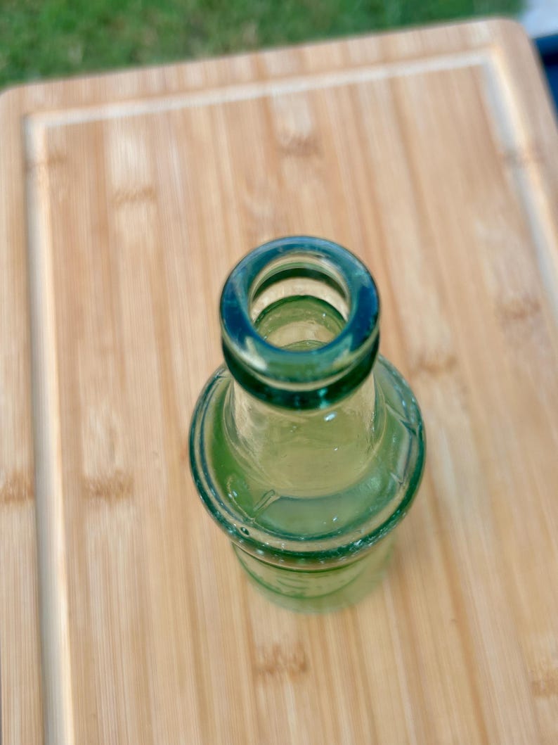 May include: A small, translucent green glass bottle viewed from directly above. The bottle features a rounded neck and a thick, circular rim opening. The glass has a smooth, reflective surface with subtle variations in the green tint throughout the body.