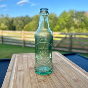 May include: A vintage light green glass bottle with a narrow neck and a rounded body. Embossed on the front is a circular design containing the text The Original 3C Drink, with additional embossed text at the base reading REG US PAT OFF.