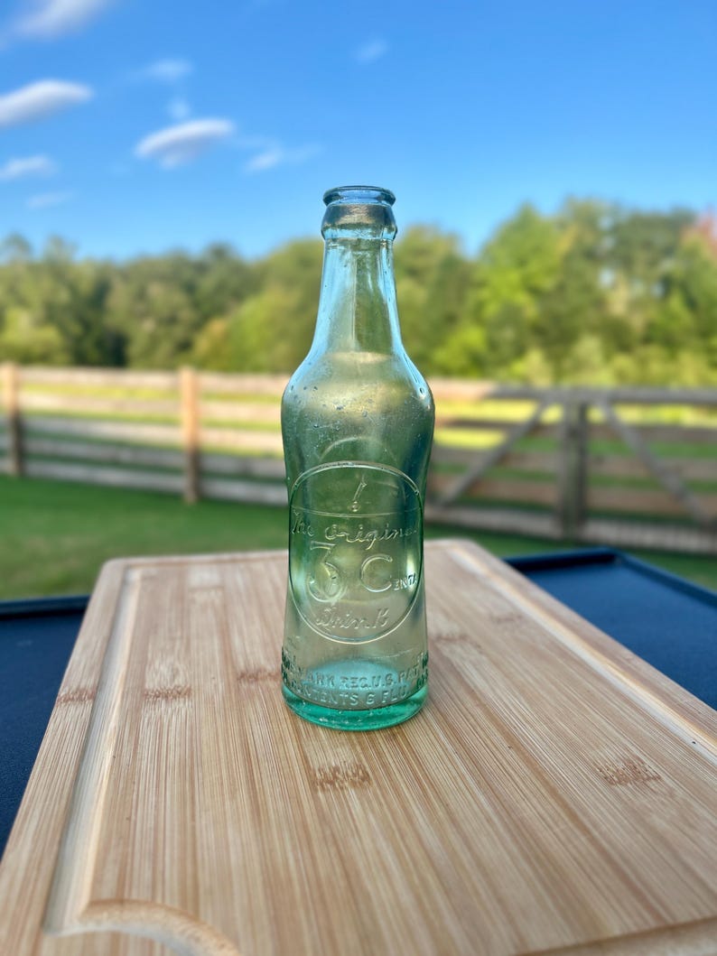 May include: A vintage light green glass bottle with a narrow neck and a rounded body. Embossed on the front is a circular design containing the text The Original 3C Drink, with additional embossed text at the base reading REG US PAT OFF.