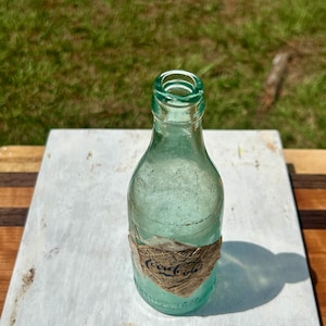 Pode incluir: Uma garrafa de vidro verde-claro vintage com gargalo estreito e corpo arredondado. Um r&oacute;tulo de papel desgastado em forma de diamante est&aacute; colado na parte inferior, com o texto azul-escuro Coca-Cola impresso em fonte cursiva cl&aacute;ssica.