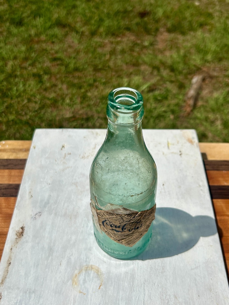 Pode incluir: Uma garrafa de vidro verde-claro vintage com gargalo estreito e corpo arredondado. Um r&oacute;tulo de papel desgastado em forma de diamante est&aacute; colado na parte inferior, com o texto azul-escuro Coca-Cola impresso em fonte cursiva cl&aacute;ssica.
