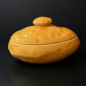 May include: A ceramic serving dish with a lid, in a warm yellow color. The dish has an oval shape with a rounded lid and a small, rounded knob on top. The dish is on a black background.