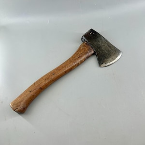 May include: A hand axe featuring a curved, weathered wooden handle with a natural grain and a dark, metallic steel head. The metal blade shows signs of wear and oxidation, with a slightly curved cutting edge attached to the wooden shaft.