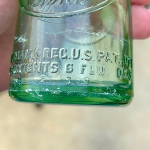 May include: A green glass bottle with embossed text that reads Drink, TRADE MARK REG. U.S. PAT. OFF., and CONTENTS 6 FLU. OZS. The bottle features a curved design with clear lettering molded directly into the glass surface, showing a capacity of 6 fluid ounces.