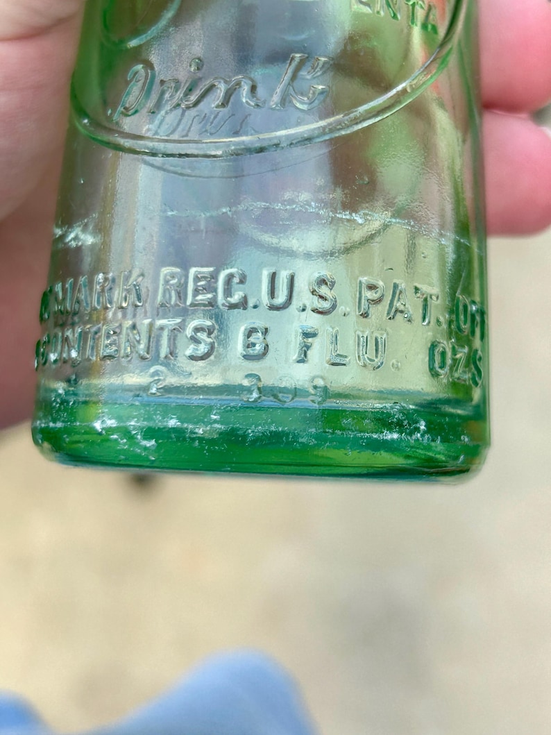 May include: A green glass bottle with embossed text that reads Drink, TRADE MARK REG. U.S. PAT. OFF., and CONTENTS 6 FLU. OZS. The bottle features a curved design with clear lettering molded directly into the glass surface, showing a capacity of 6 fluid ounces.