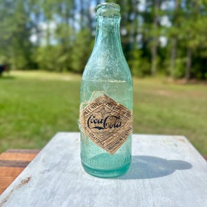Pode incluir: Uma garrafa de vidro verde-claro vintage da Coca-Cola com um r&oacute;tulo de papel em forma de diamante. O r&oacute;tulo apresenta o texto Coca-Cola, Authorized under the authority of the Coca-Cola Co. Atlanta GA e Minimum Volume. Birmingham ALA est&aacute; gravado.