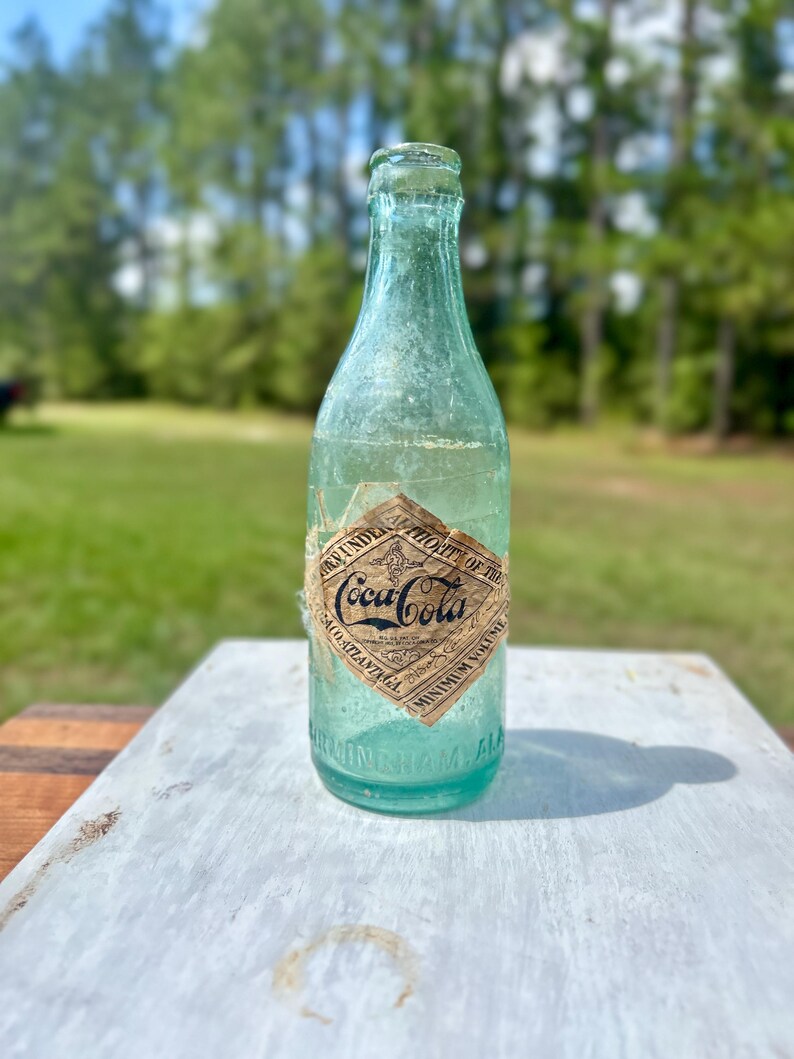 Pode incluir: Uma garrafa de vidro verde-claro vintage da Coca-Cola com um r&oacute;tulo de papel em forma de diamante. O r&oacute;tulo apresenta o texto Coca-Cola, Authorized under the authority of the Coca-Cola Co. Atlanta GA e Minimum Volume. Birmingham ALA est&aacute; gravado.