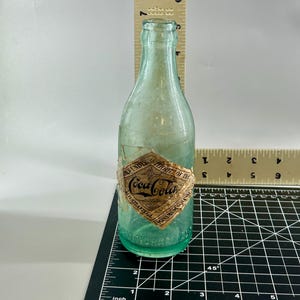 Pode incluir: Uma garrafa de vidro verde claro vintage da Coca-Cola com um r&oacute;tulo de papel em forma de diamante escrito Coca-Cola, Authority of the Coca-Cola Co. Atlanta GA. Uma r&eacute;gua mostra que tem cerca de 19,7 cm de altura e 6,4 cm de largura.
