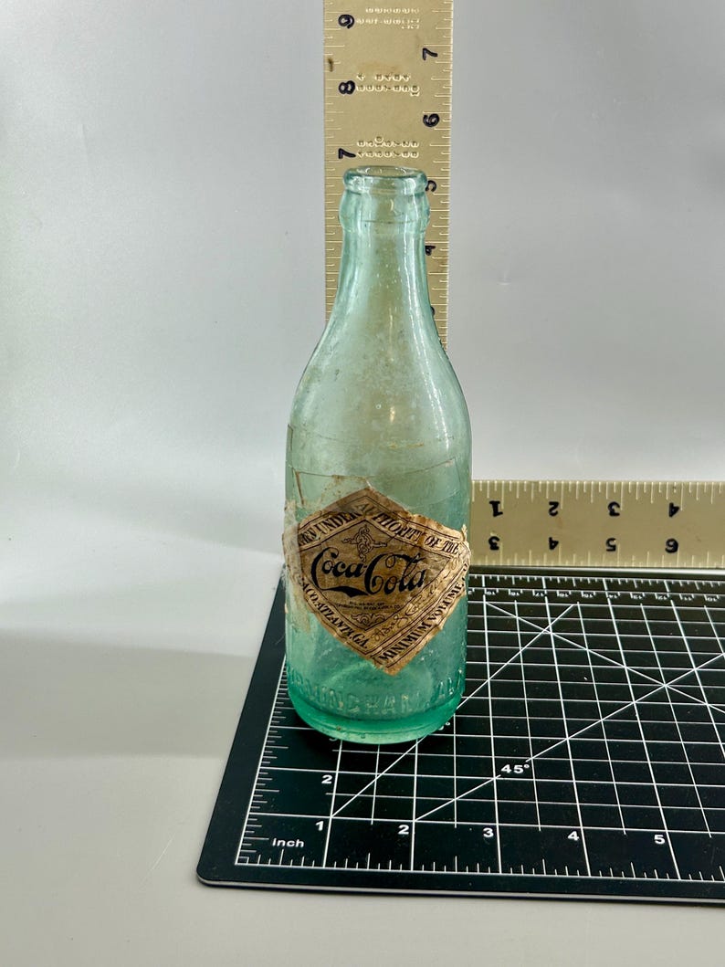 Pode incluir: Uma garrafa de vidro verde claro vintage da Coca-Cola com um r&oacute;tulo de papel em forma de diamante escrito Coca-Cola, Authority of the Coca-Cola Co. Atlanta GA. Uma r&eacute;gua mostra que tem cerca de 19,7 cm de altura e 6,4 cm de largura.