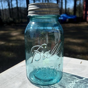May include: A vintage blue Ball Perfect Mason jar with a metal lid. The jar has embossed lettering that reads "Ball" and "Perfect Mason". The glass is a vibrant blue color, and the lid is a tarnished silver. The jar is sitting on a light-colored surface.