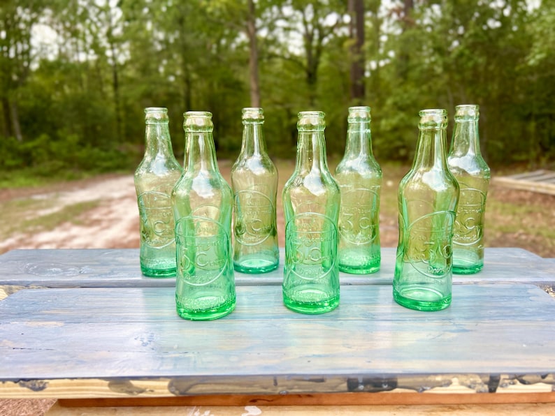 Group of seven aqua glass 3 Centa soda bottles arranged outdoors on a white surface.