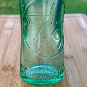 May include: A vintage light green glass bottle with embossed text. The center features a circular design with the text The Original 3 Cent Drink. The base includes embossed text reading TRADE MARK REG U.S. PAT. OFF. MIN. CONTENTS 6 FL. OZ.