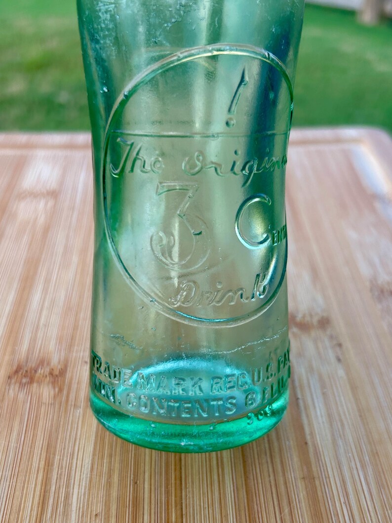 May include: A vintage light green glass bottle with embossed text. The center features a circular design with the text The Original 3 Cent Drink. The base includes embossed text reading TRADE MARK REG U.S. PAT. OFF. MIN. CONTENTS 6 FL. OZ.