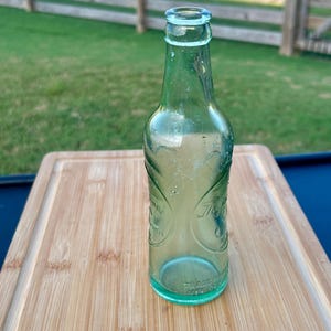 May include: A vintage light green glass soda bottle with a narrow neck and a rounded rim. The bottle features embossed decorative scrollwork patterns and circular text elements on the body, with the words TRADE MARK embossed near the base.