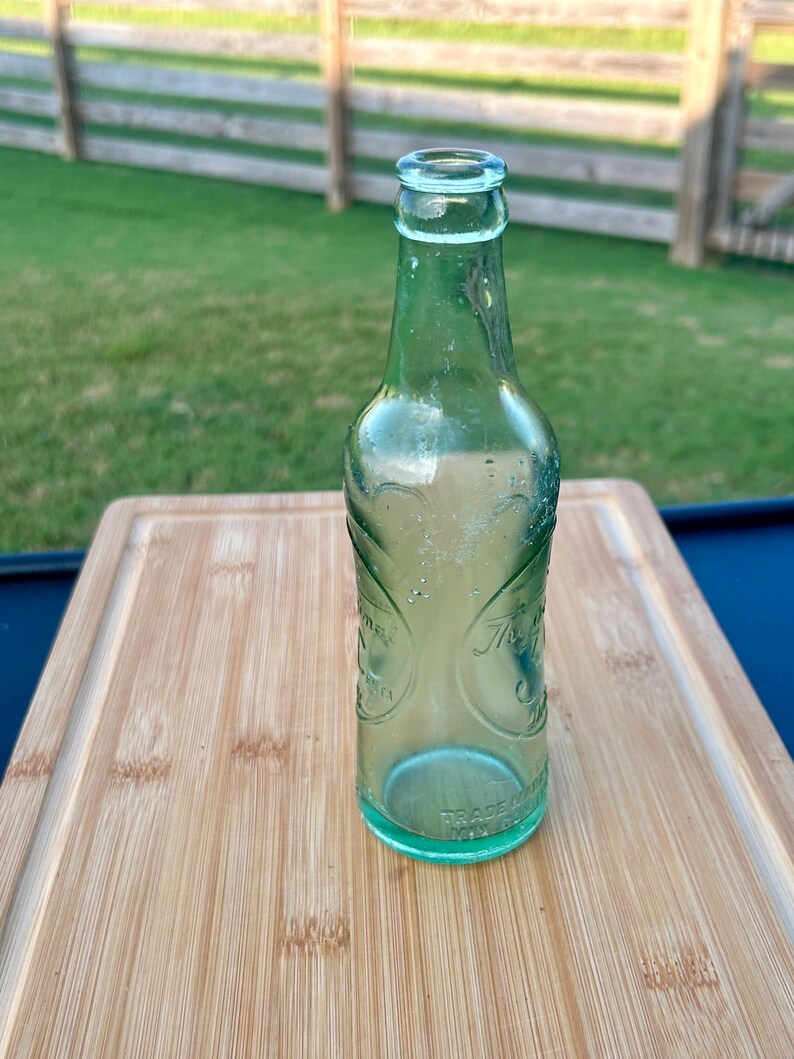 May include: A vintage light green glass soda bottle with a narrow neck and a rounded rim. The bottle features embossed decorative scrollwork patterns and circular text elements on the body, with the words TRADE MARK embossed near the base.
