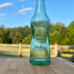 May include: A vintage light green glass soda bottle with a narrow neck and a rounded base. Embossed on the front is a circular logo containing the text The Original 3 C Drink, featuring a stylized letter C and a small graphic of a globe.