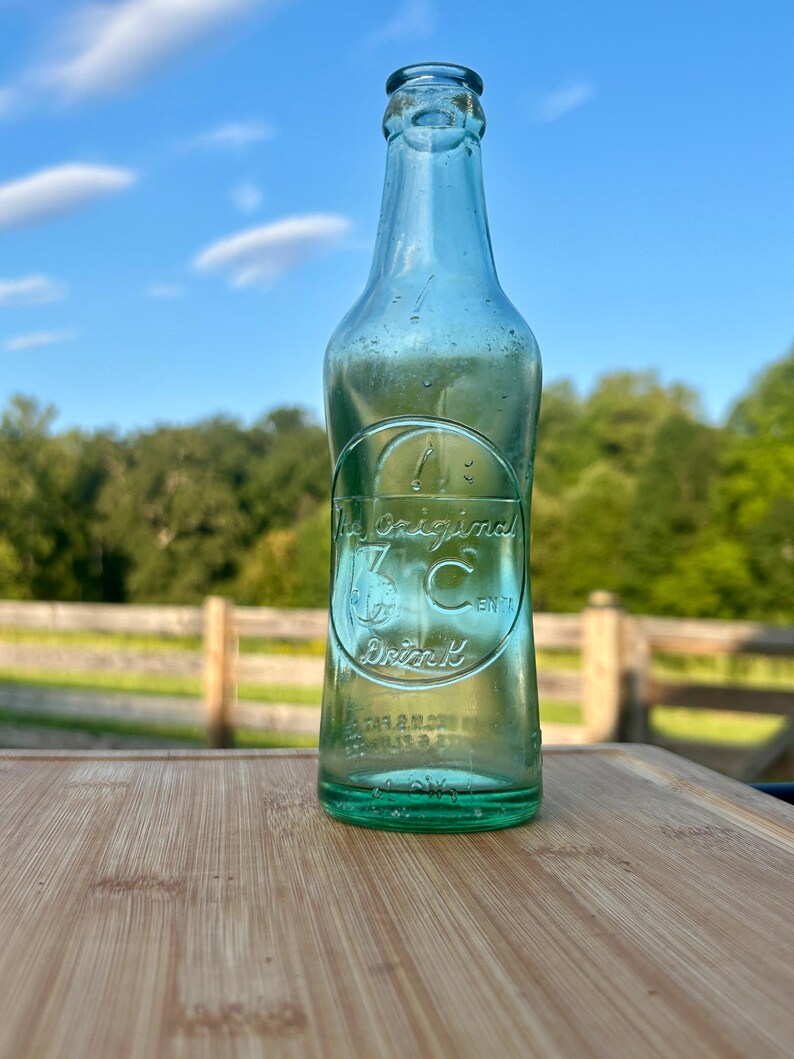 May include: A vintage light green glass soda bottle with a narrow neck and a rounded base. Embossed on the front is a circular logo containing the text The Original 3 C Drink, featuring a stylized letter C and a small graphic of a globe.