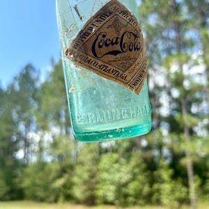 Pode incluir: Uma garrafa de vidro Coca-Cola azul-claro vintage com um r&oacute;tulo de papel em forma de diamante com o texto preto BOTTLED UNDER AUTHORITY OF COCA-COLA CO. ATLANTA GA. A palavra BIRMINGHAM est&aacute; gravada em letras mai&uacute;sculas na base de vidro.