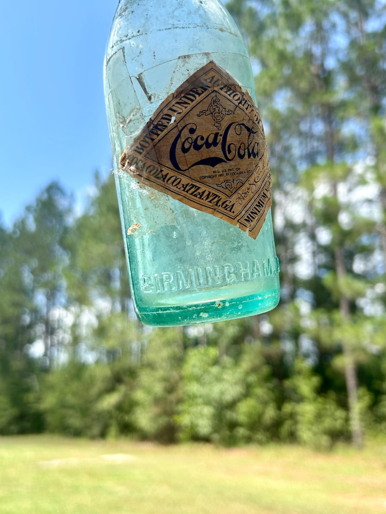Pode incluir: Uma garrafa de vidro Coca-Cola azul-claro vintage com um r&oacute;tulo de papel em forma de diamante com o texto preto BOTTLED UNDER AUTHORITY OF COCA-COLA CO. ATLANTA GA. A palavra BIRMINGHAM est&aacute; gravada em letras mai&uacute;sculas na base de vidro.