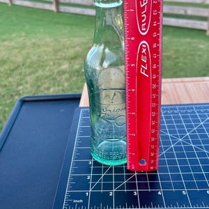May include: A clear glass bottle with a narrow neck and embossed text reading The Original 7up Drink. A red Flexi Ruler is placed vertically against the bottle, showing a height of approximately 8 inches from the base to the top of the bottle opening.