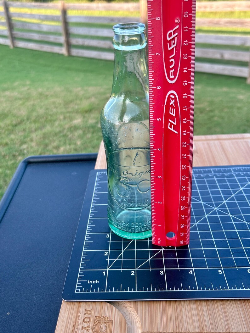 May include: A clear glass bottle with a narrow neck and embossed text reading The Original 7up Drink. A red Flexi Ruler is placed vertically against the bottle, showing a height of approximately 8 inches from the base to the top of the bottle opening.
