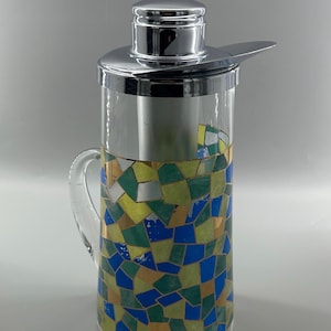 May include: A clear glass cocktail shaker with a chrome lid and handle. The lower section features a mosaic pattern of blue, green, yellow, and gold tiles. The lid includes a strainer. A stylish barware item.