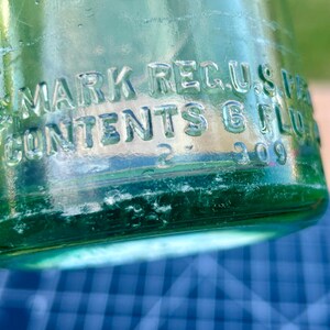 May include: A light green glass bottle with embossed text reading MARK REG. U.S. PAT. OFF. and CONTENTS 6 FL. OZ. The base of the bottle features the numbers 2 and 309 embossed into the glass surface, showing clear molded lettering and details.