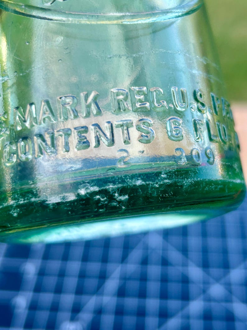 May include: A light green glass bottle with embossed text reading MARK REG. U.S. PAT. OFF. and CONTENTS 6 FL. OZ. The base of the bottle features the numbers 2 and 309 embossed into the glass surface, showing clear molded lettering and details.