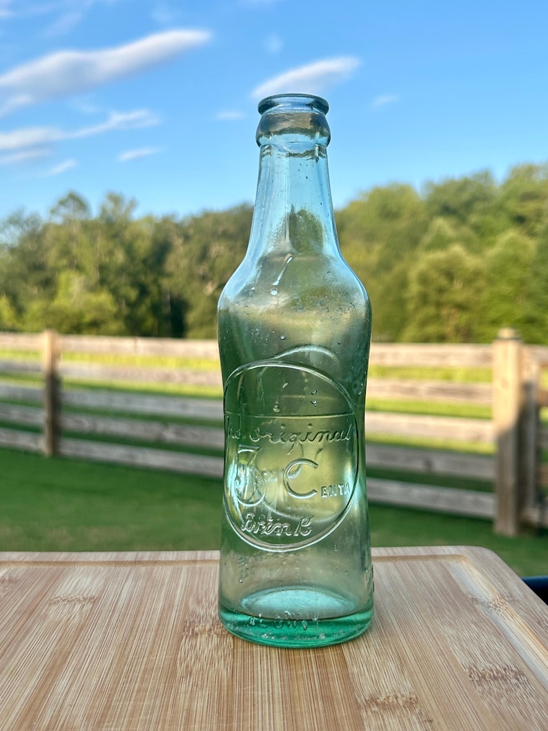 May include: A clear, light green glass bottle with an embossed circular design on the front. The text inside the circle reads The Original 5 Cent Drink. The bottle has a narrow neck and a rounded rim, featuring a smooth, transparent glass surface.