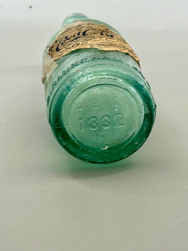 Pode incluir: Uma garrafa de vidro verde claro vintage com um r&oacute;tulo de papel desgastado apresentando o texto Coca-Cola. A base da garrafa &eacute; gravada com as letras A B CO e o n&uacute;mero 1332. O vidro tem um tom de &aacute;gua distinto e fundo arredondado.