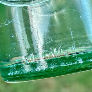 May include: A translucent emerald green glass bottle featuring embossed lettering. The upper portion displays the word Drink in a curved script, while the lower section features the embossed text al G.W. on the smooth, rounded glass surface.