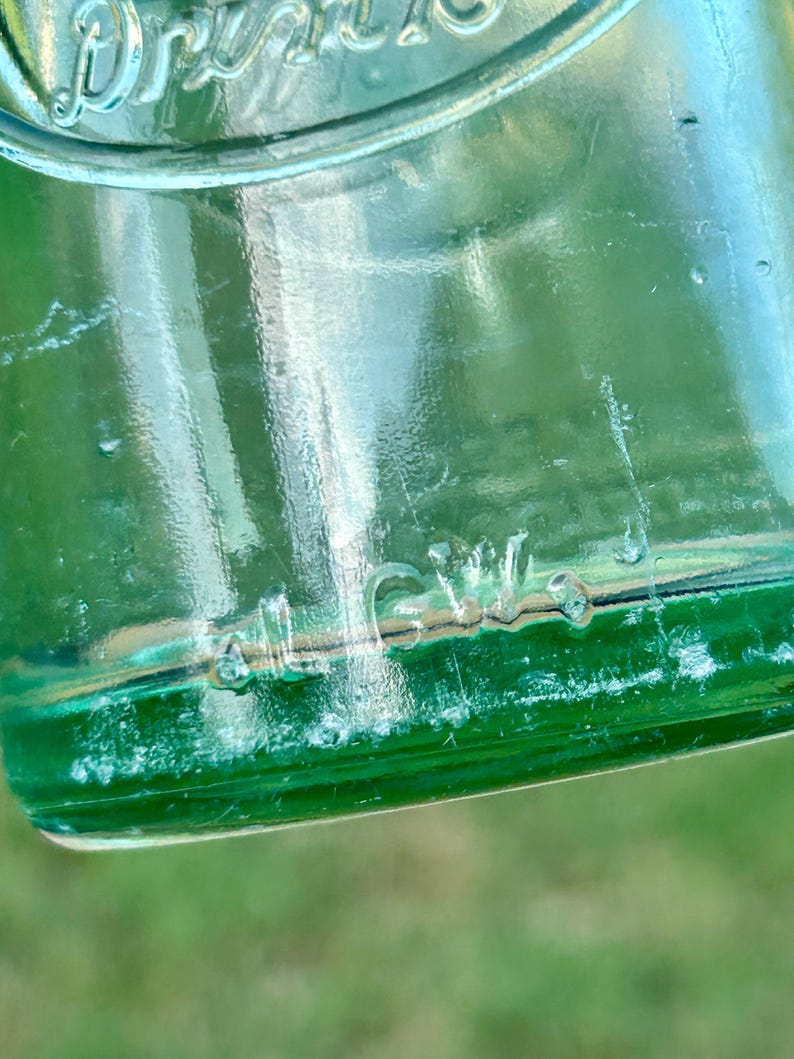 May include: A translucent emerald green glass bottle featuring embossed lettering. The upper portion displays the word Drink in a curved script, while the lower section features the embossed text al G.W. on the smooth, rounded glass surface.